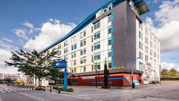 A modern hotel building with a prominent 'aloft' sign, multiple floors, and a clear sky.