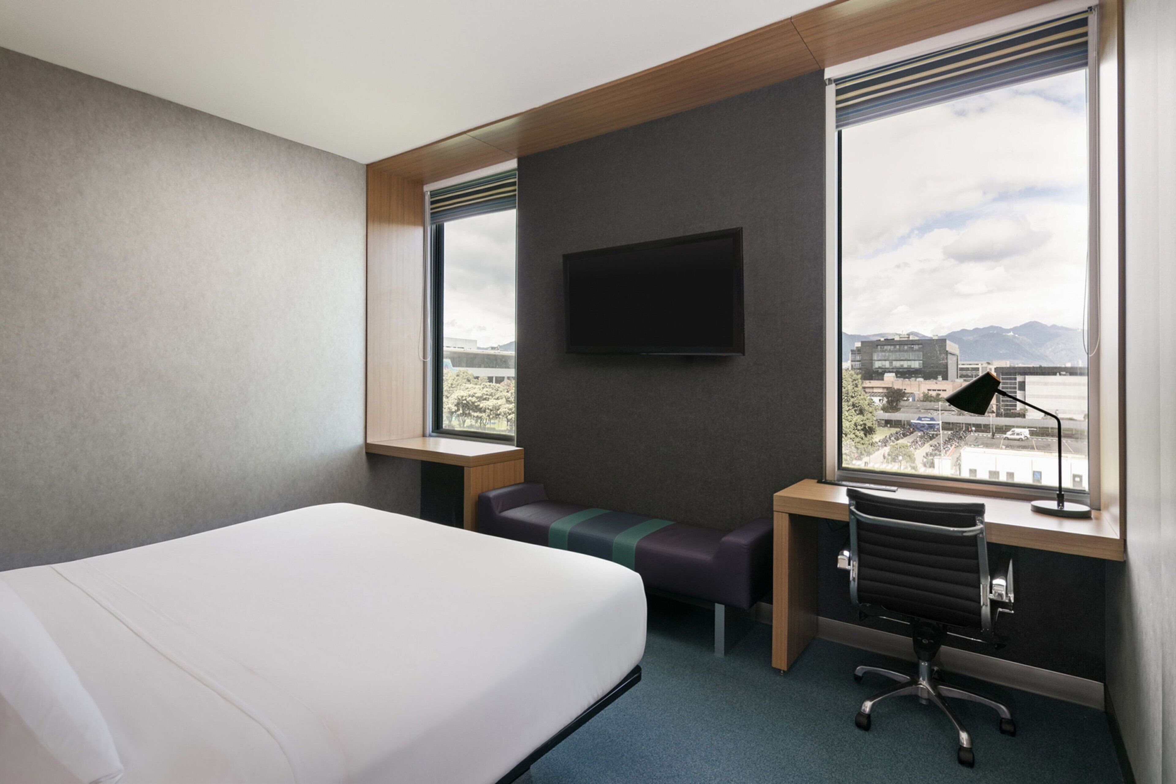 Quarto de hotel com uma cama, uma escrivaninha, uma cadeira, televisão e janela com vista para a cidade.