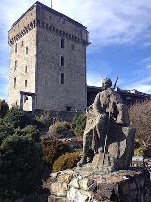 Una estatua de piedra de una figura sentada con un bastón, una torre del reloj al fondo y un letrero que indica una terraza.