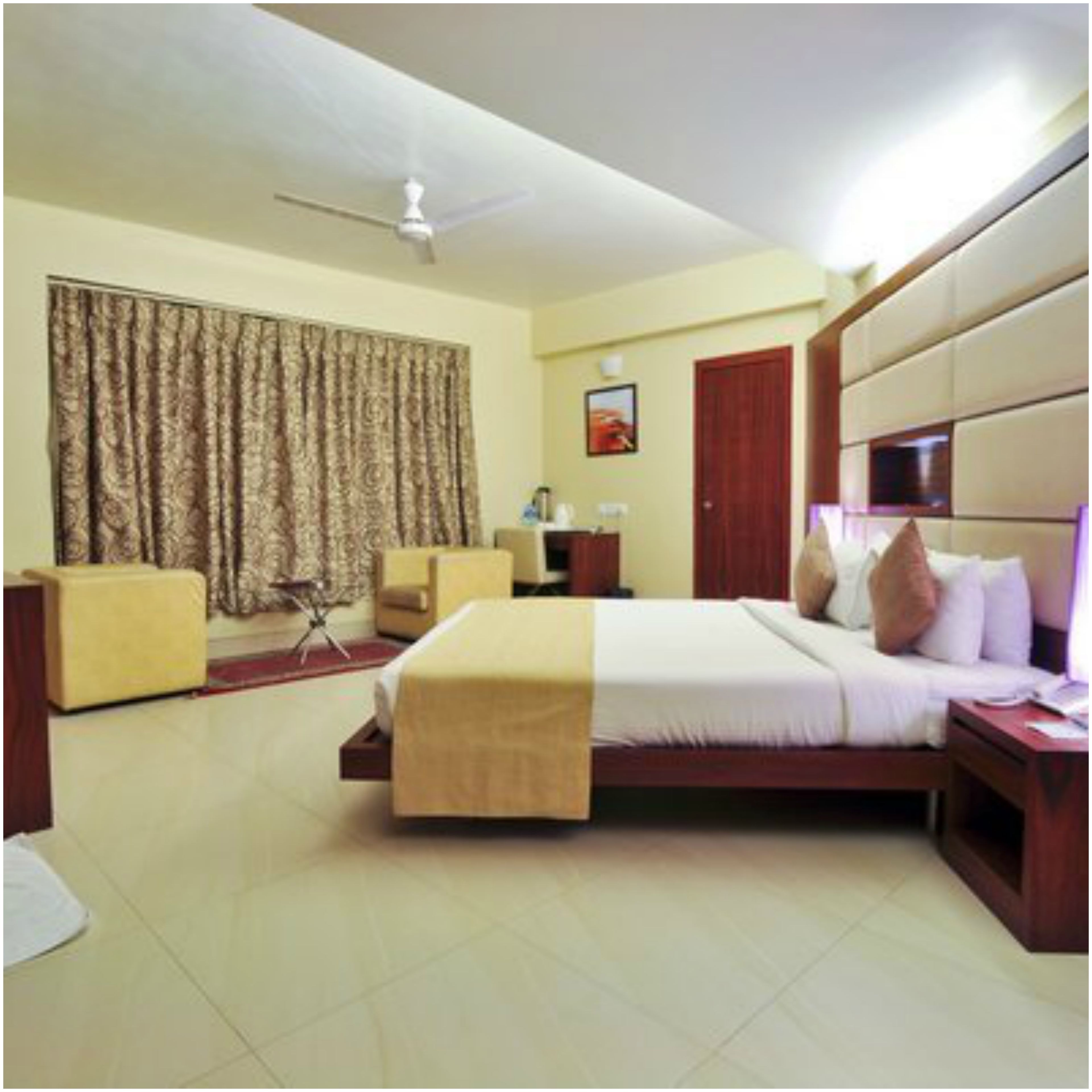 A hotel room with a bed, a sofa, a desk, a chair, a curtain, a ceiling fan, and a picture on the wall.