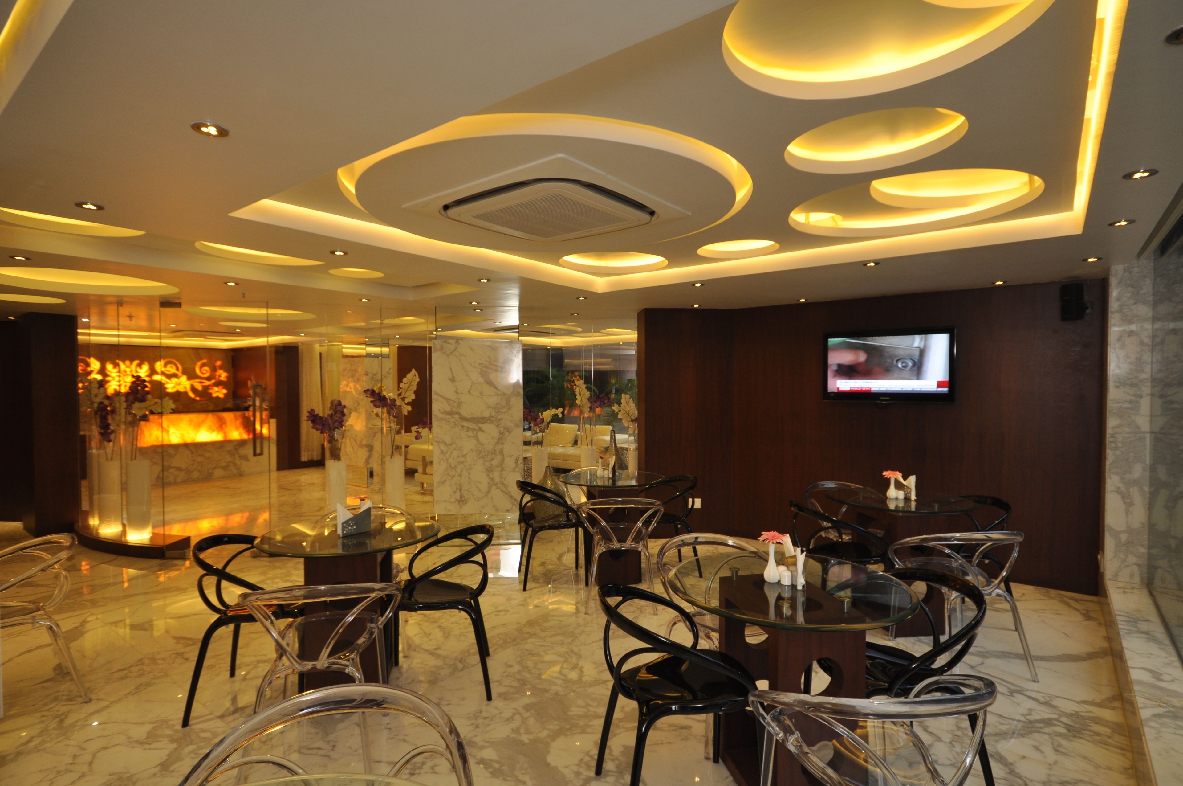 A modern hotel lobby with a marble floor, glass tables, and black chairs. There is a reception area with a fire feature and a TV mounted on the wall.