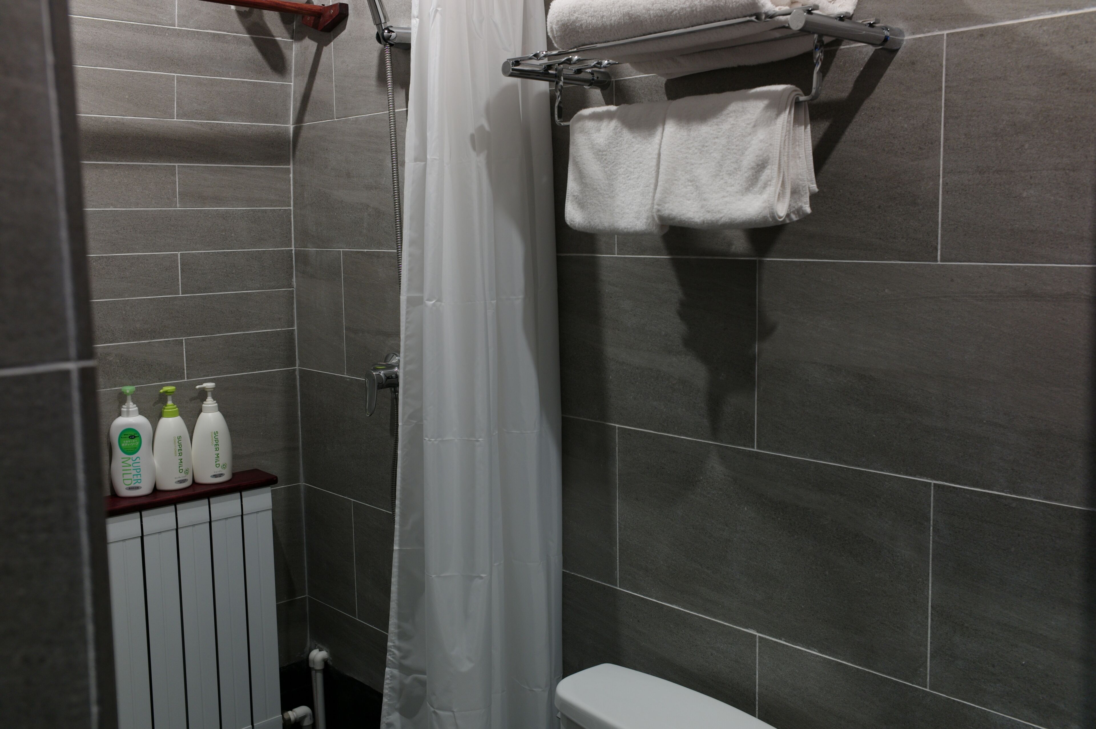 A modern bathroom with grey tiles, a shower curtain, a towel rack, and three bottles of toiletries on a shelf.