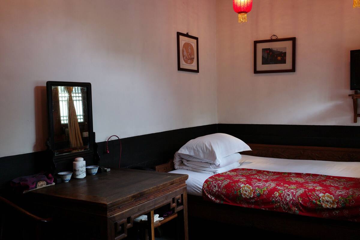 A room with a wooden desk, a bed with a red floral bedspread, and two framed pictures on the wall.