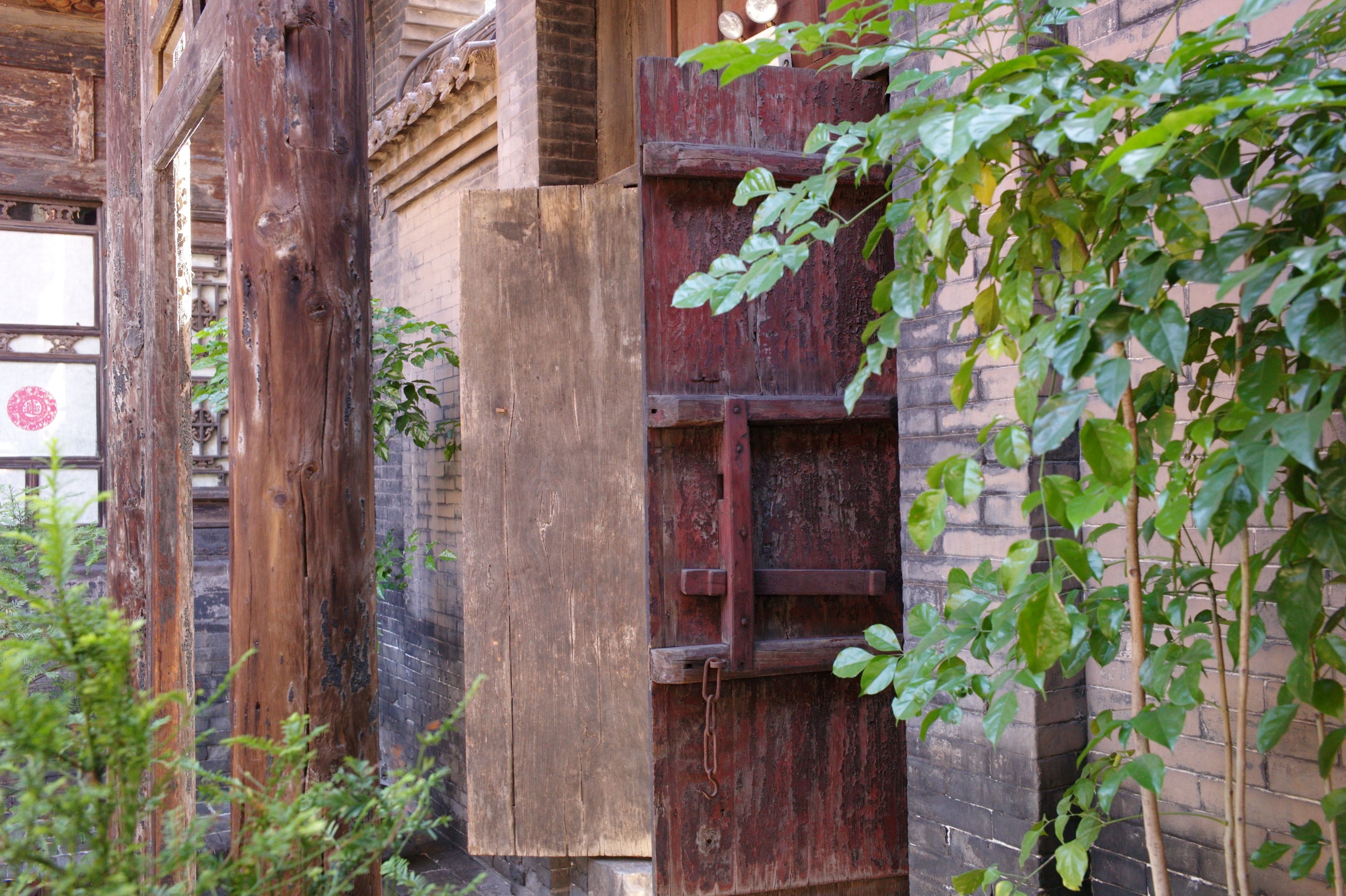 Une porte en bois rustique, dotée d’un loquet en métal, entourée de verdure et bordée d’un mur de briques.