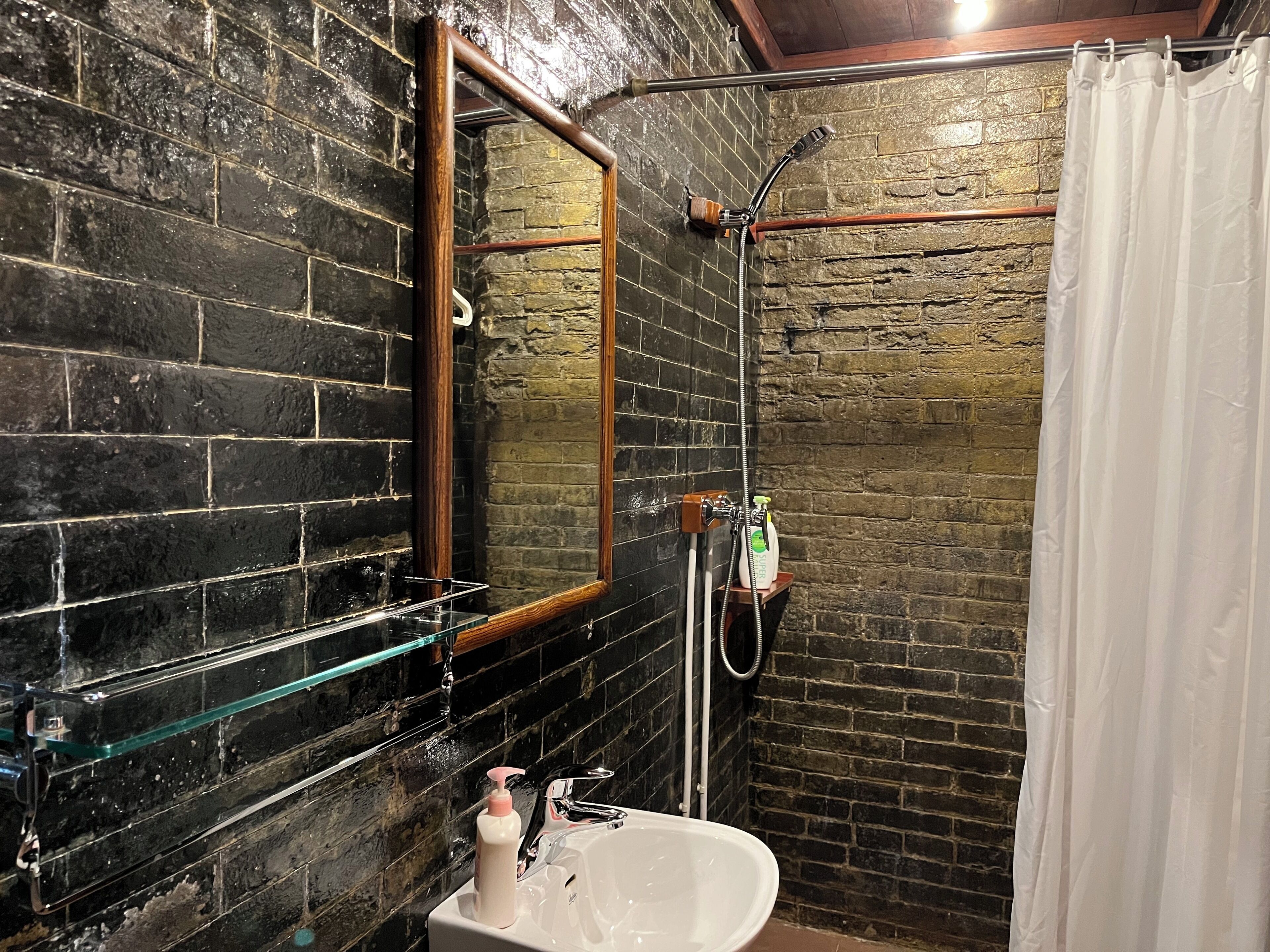 A bathroom with a white sink, a glass shelf, a wooden-framed mirror, a shower with a hose, and a brick wall.