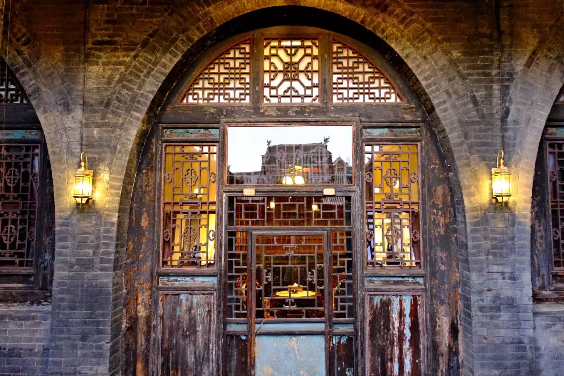 Une porte en arc surmontée d’un motif traditionnel chinois en forme de treillis, une lanterne accrochée au mur et, au loin, on aperçoit un bâtiment.