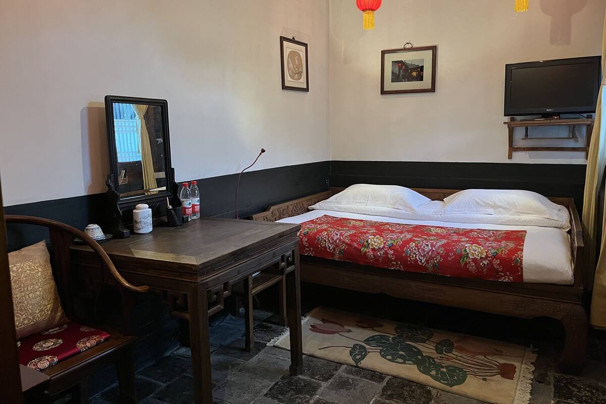A hotel room with a bed, a wooden table, a chair, a television, and two red lanterns.