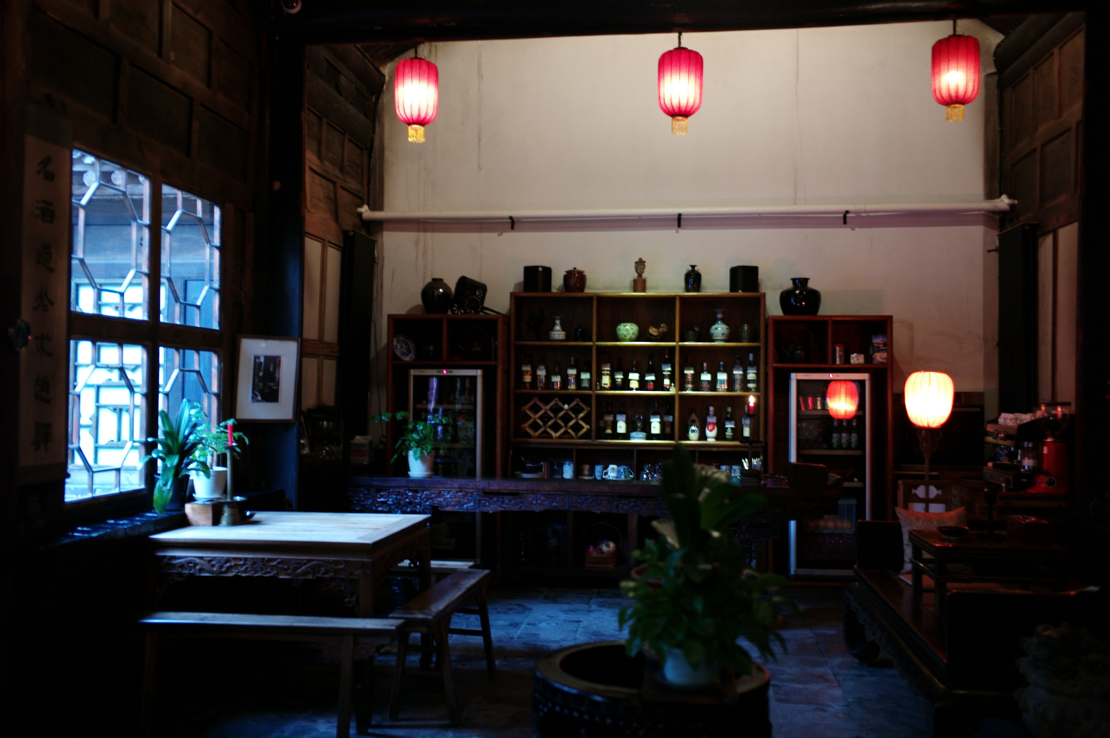 A dimly lit room with wooden furniture, a bar area with shelves of bottles, and traditional lanterns.
