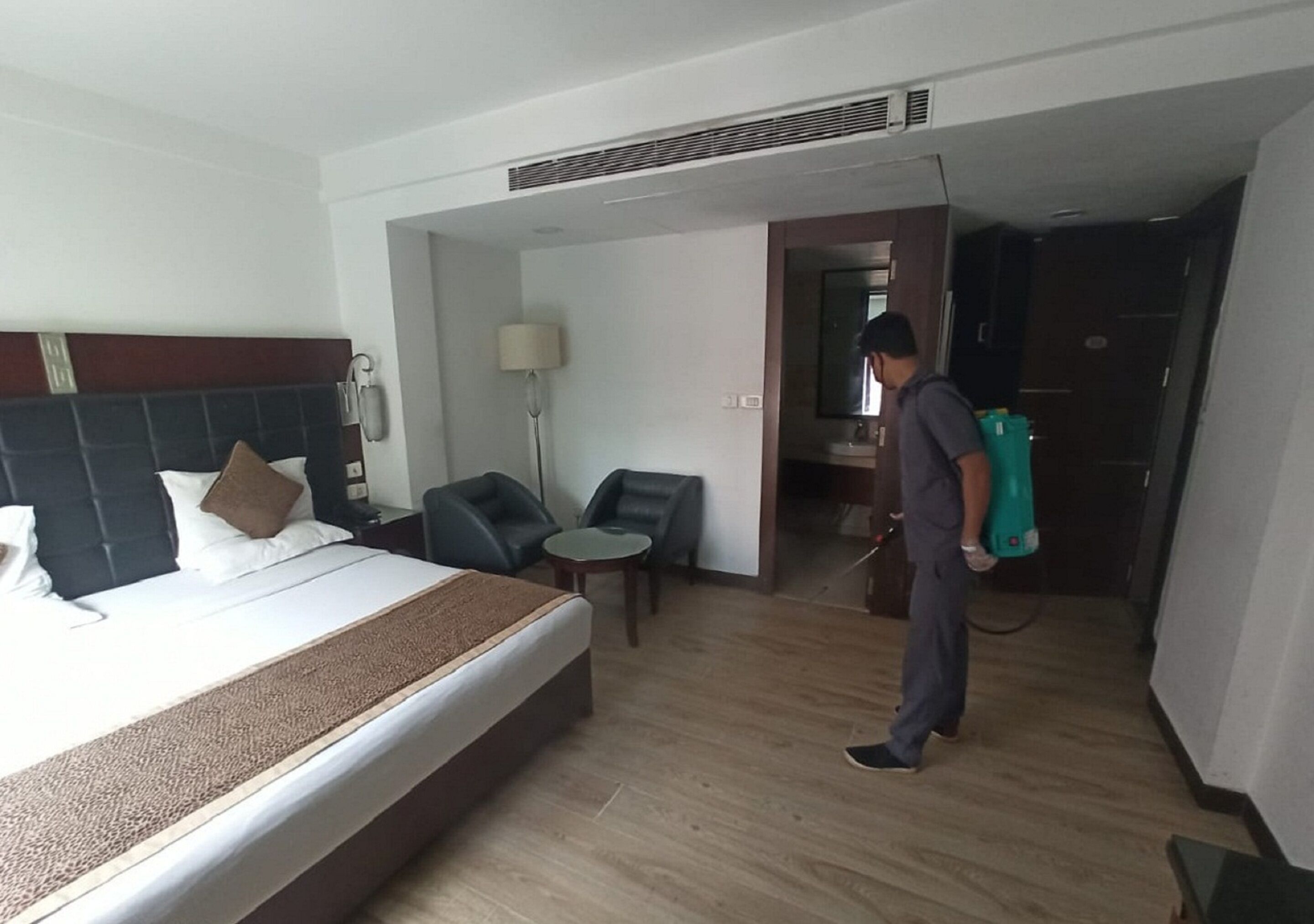 A hotel room with a bed, a nightstand, a lamp, a chair, and a person using a cleaning device.