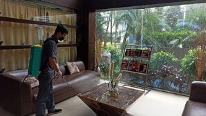 Une personne est en train de nettoyer un salon à l’aide d’un aspirateur. La pièce est dotée d’une grande fenêtre donnant sur un paysage verdoyant. On y trouve un canapé en cuir marron et une table basse en verre sur laquelle est posé un vase.