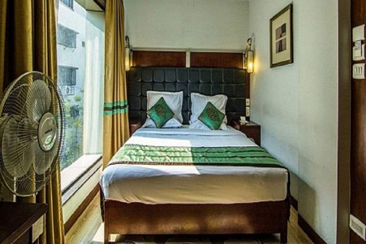 A hotel room with a bed, a nightstand, a wall-mounted lamp, a window with curtains, and a standing fan.