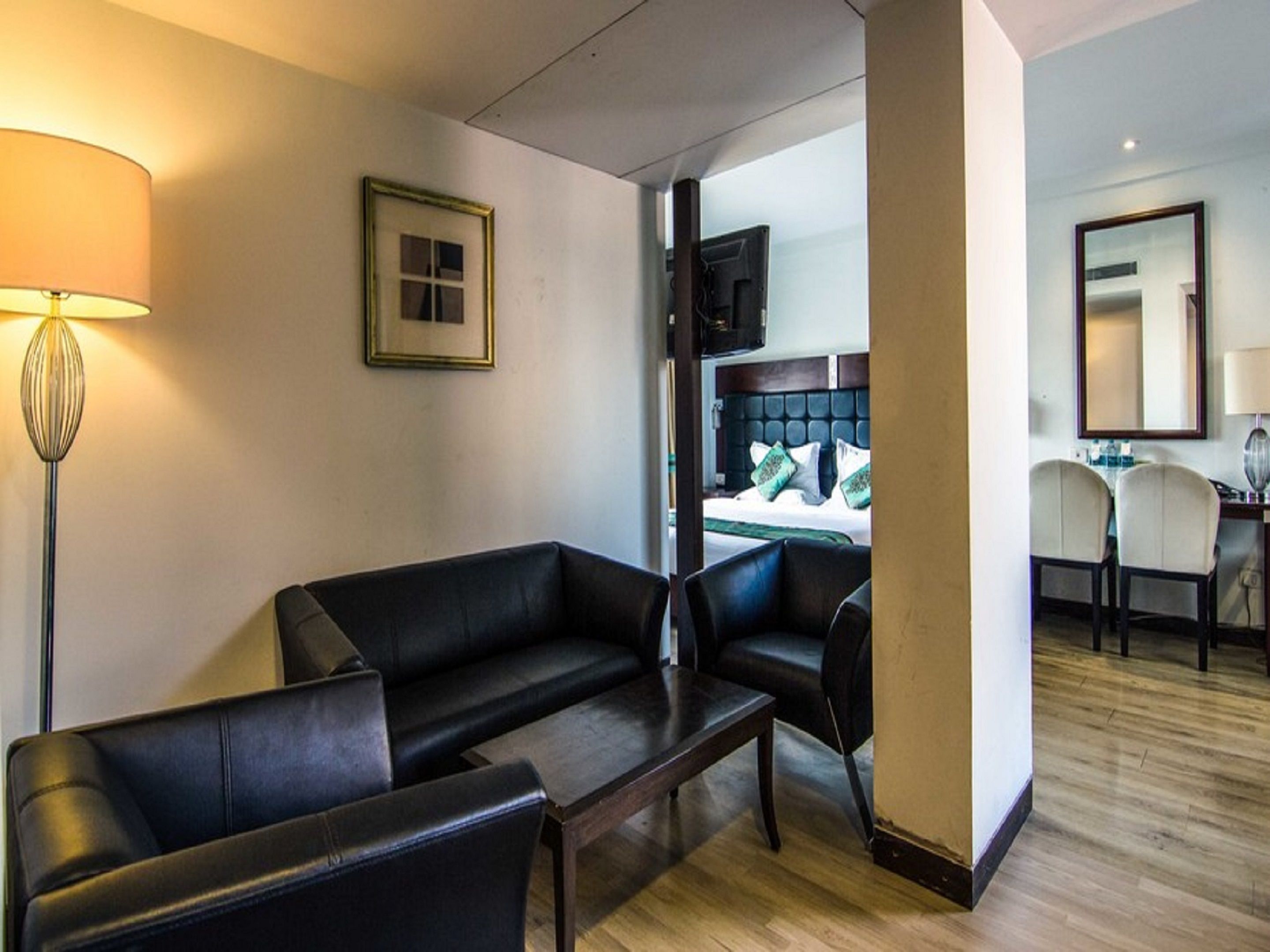 Une chambre d’hôtel avec un canapé en cuir noir, une table basse en bois et un bureau avec deux chaises. On aperçoit un lit avec des oreillers verts à travers une porte ouverte.
