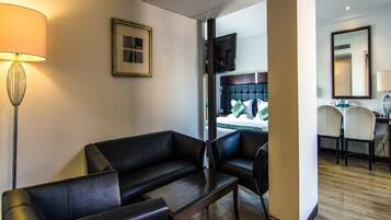 Une chambre d’hôtel avec un canapé en cuir noir, une table basse en bois et un bureau avec deux chaises. On aperçoit un lit avec des oreillers verts à travers une porte ouverte.