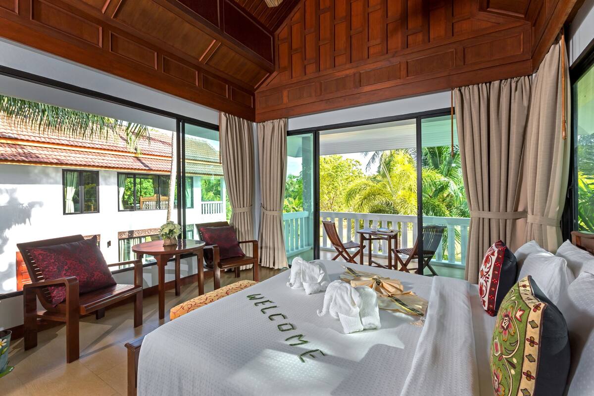 A hotel room with a large bed, a balcony with a view of the greenery, wooden furniture, and a 'WELCOME' sign on the bed.