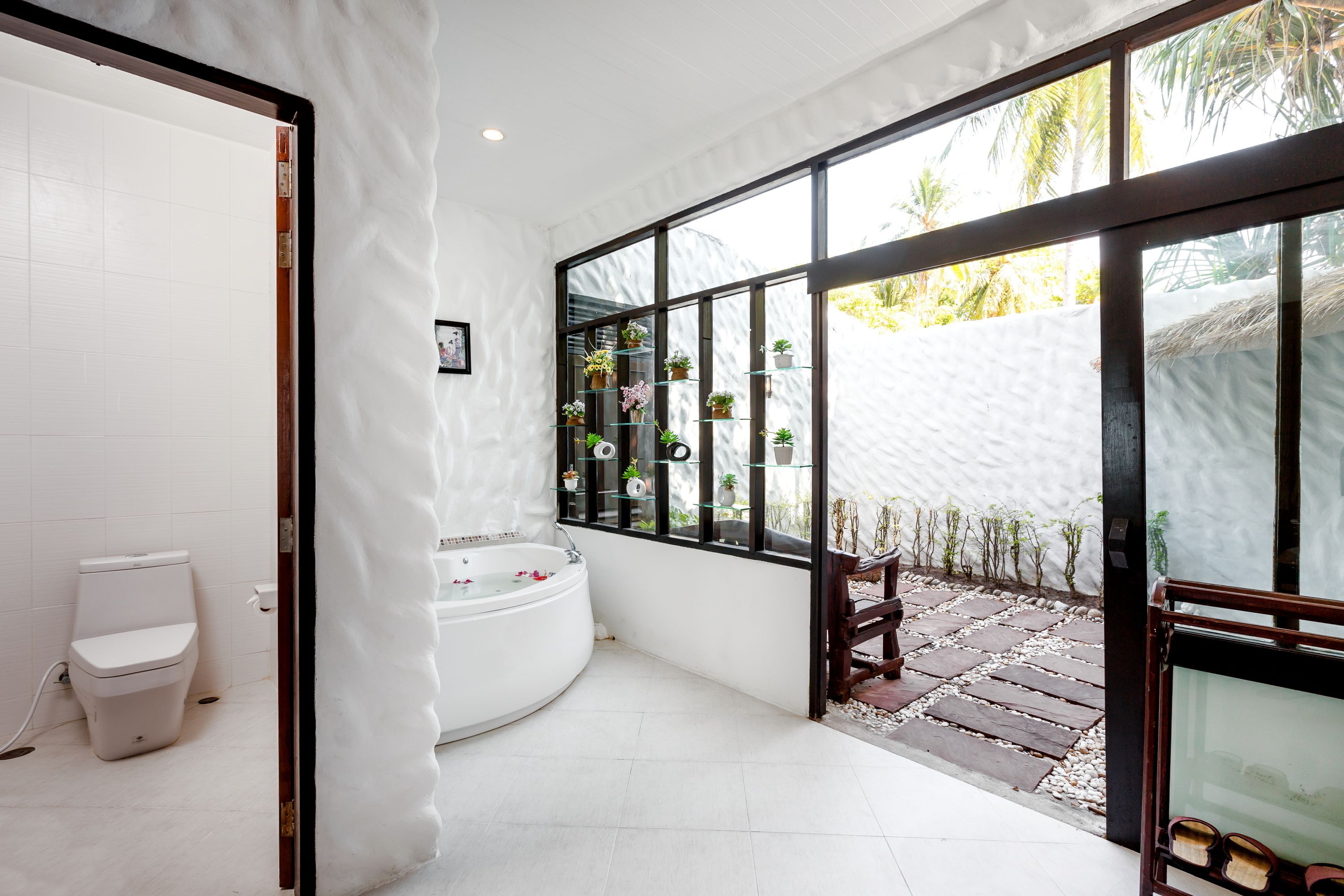A modern bathroom with a white toilet, a large bathtub, and a glass-enclosed window with a view of a garden.