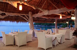 An outdoor dining area with wicker chairs and tables, set against a backdrop of a beach and ocean.