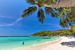 A beach with clear turquoise water, a boat on the shore, and a person standing in the water.
