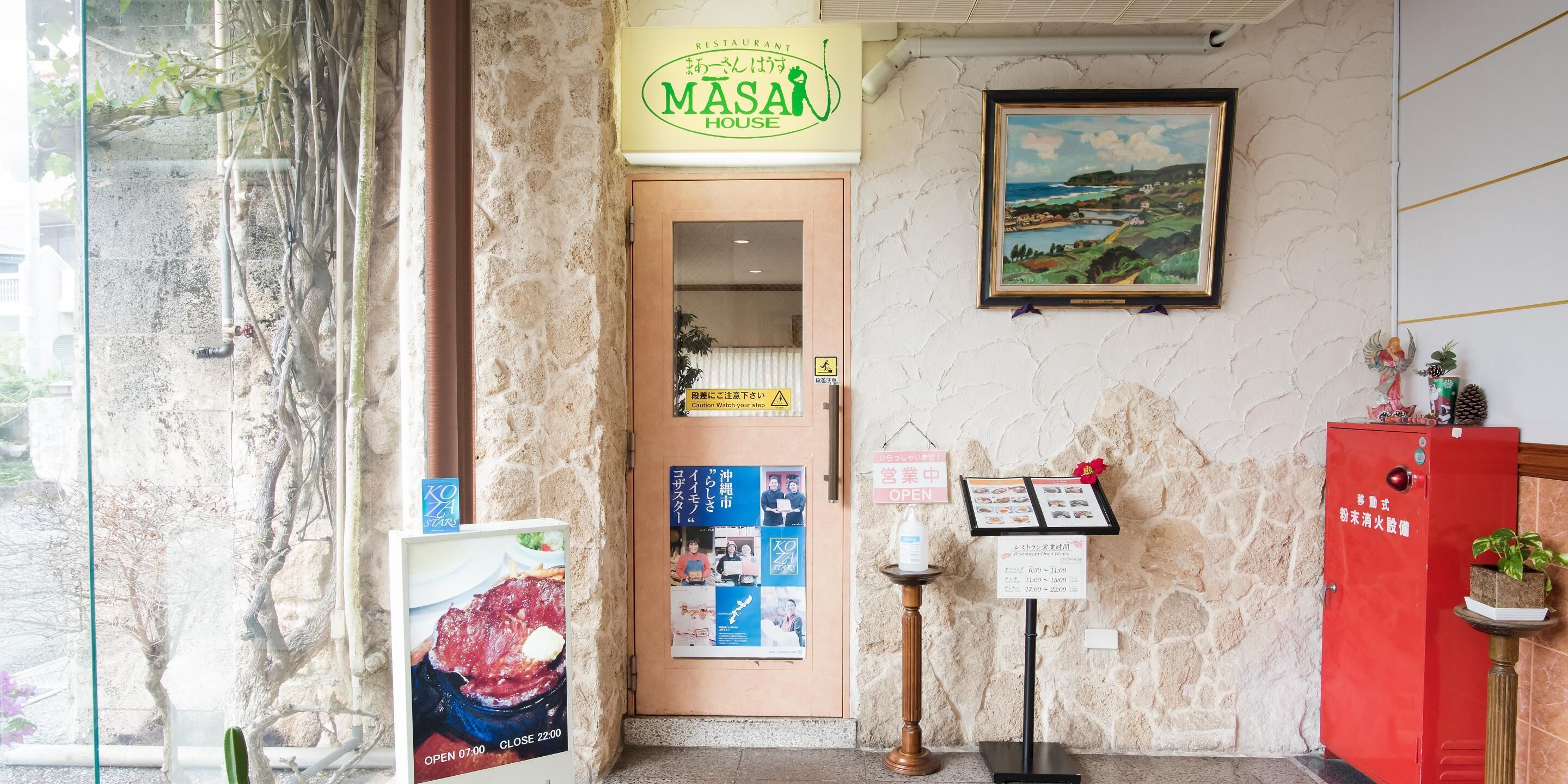 L’entrée d’un restaurant, avec une enseigne indiquant « MASAI HOUSE », un tableau de paysage encadré et divers affiches d’information.