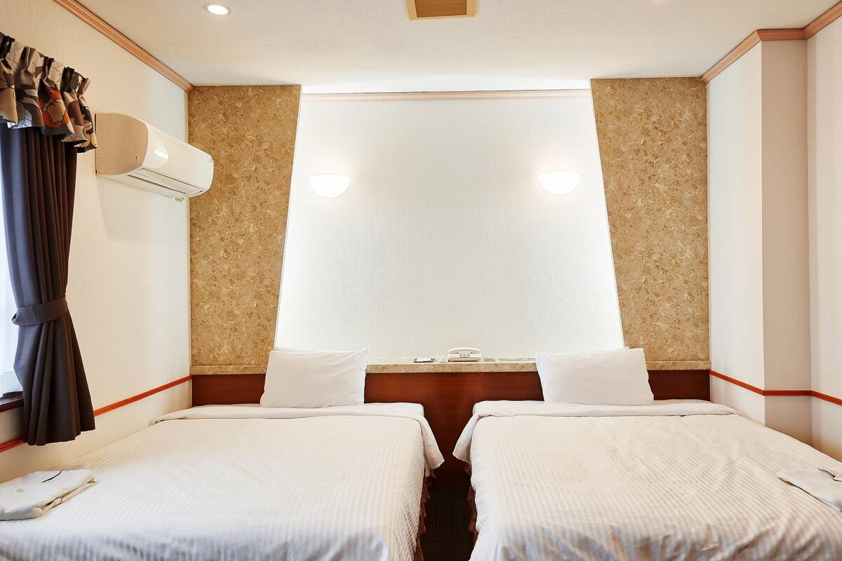 A hotel room with two beds, a window with curtains, an air conditioning unit, and a wall-mounted phone.