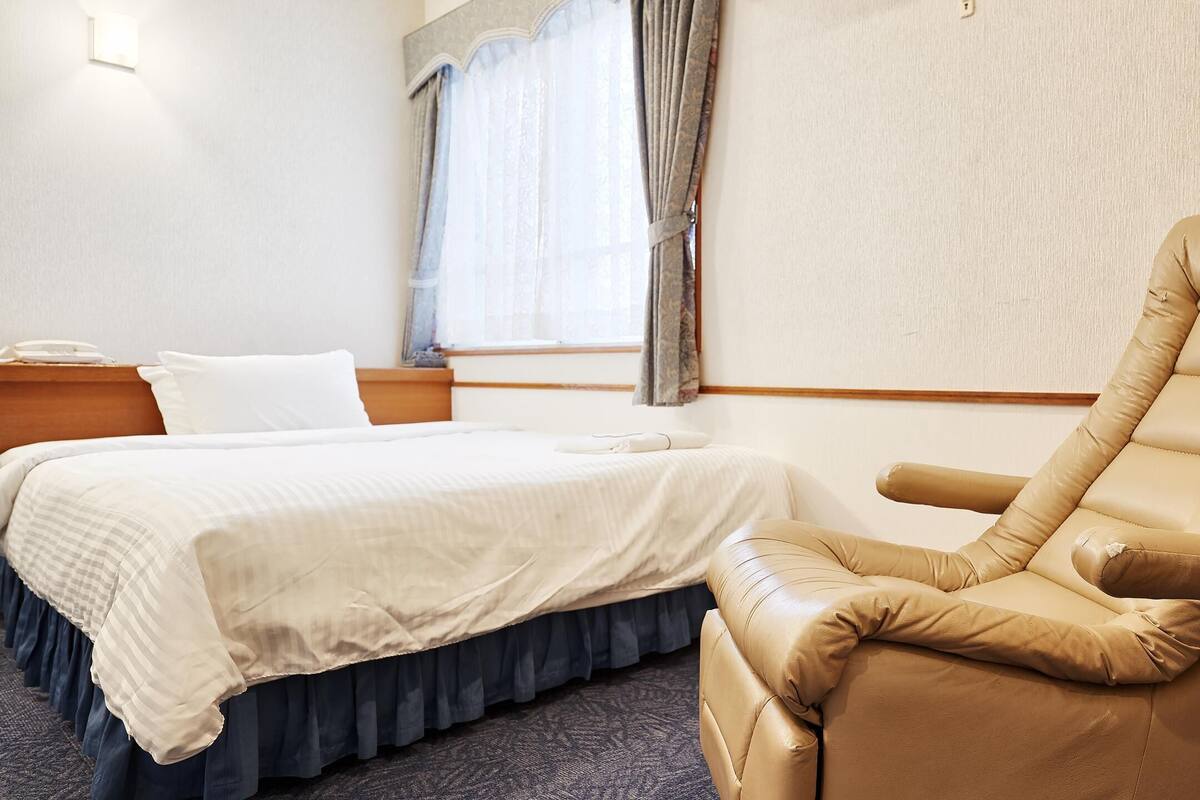 A hotel room with a bed, a reclining chair, a bedside table, and a window with curtains.