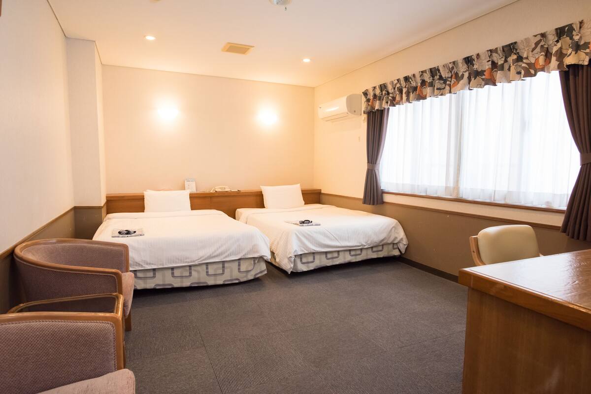A hotel room with two beds, a desk, a chair, and a window with curtains.