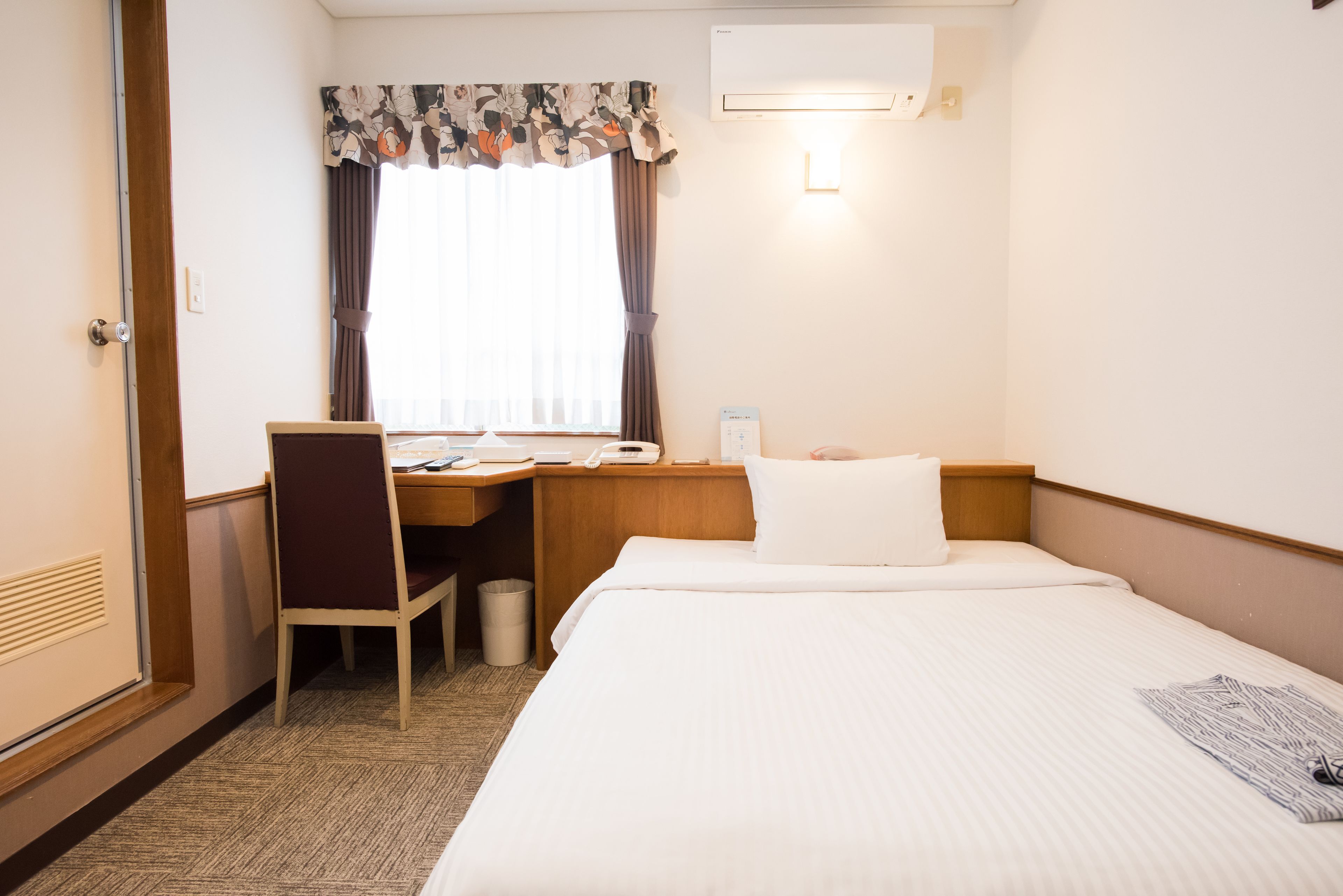 A hotel room with a bed, a desk with a chair, a waste bin, a window with curtains, and an air conditioning unit.