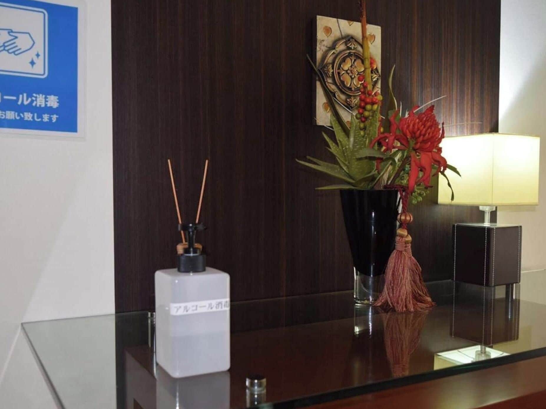 Un comptoir d’accueil avec un plateau en verre, un vase décoratif contenant des fleurs rouges, une petite lampe et une enseigne fixée au mur avec du texte en japonais.