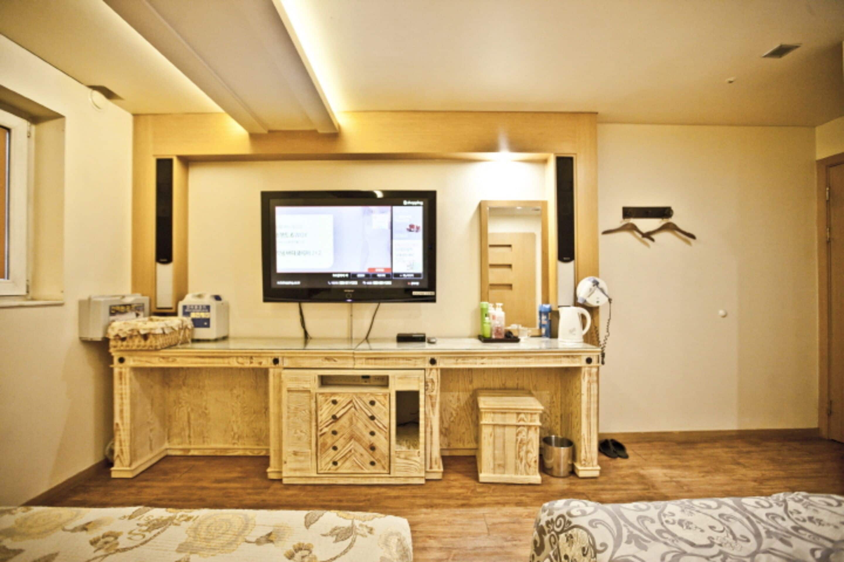 A room with a wooden desk, a television mounted on the wall, a small cabinet, and a bed with patterned bedding.