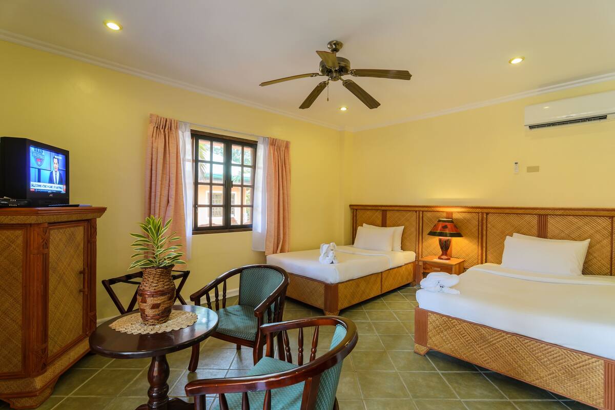 A hotel room with two single beds, a wooden headboard, a small table with chairs, a television, and a ceiling fan.