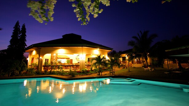 Vista nocturna de un complejo turístico con piscina, un área al aire libre cubierta con asientos y palmeras.