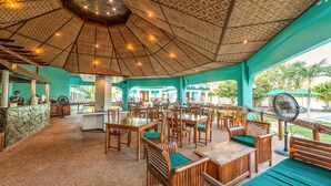 Un amplio espacio de comedor al aire libre con mesas y sillas de madera, techo de paja y vistas a la vegetación circundante.