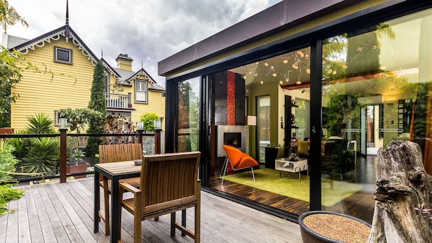 A modern house with a deck, a dining area with a table and chairs, and a large glass door leading to an interior space with a fireplace and furniture. - Corinda Contemporary (Glebe)