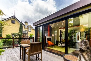 A modern house with a deck, a dining area with a table and chairs, and a large glass door leading to an interior space with a fireplace and furniture. - Corinda Contemporary (Glebe)