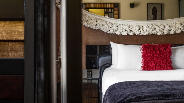 Habitación de hotel con cama, un cojín rojo, un espejo sobre la cama y un panel de madera en la pared.
