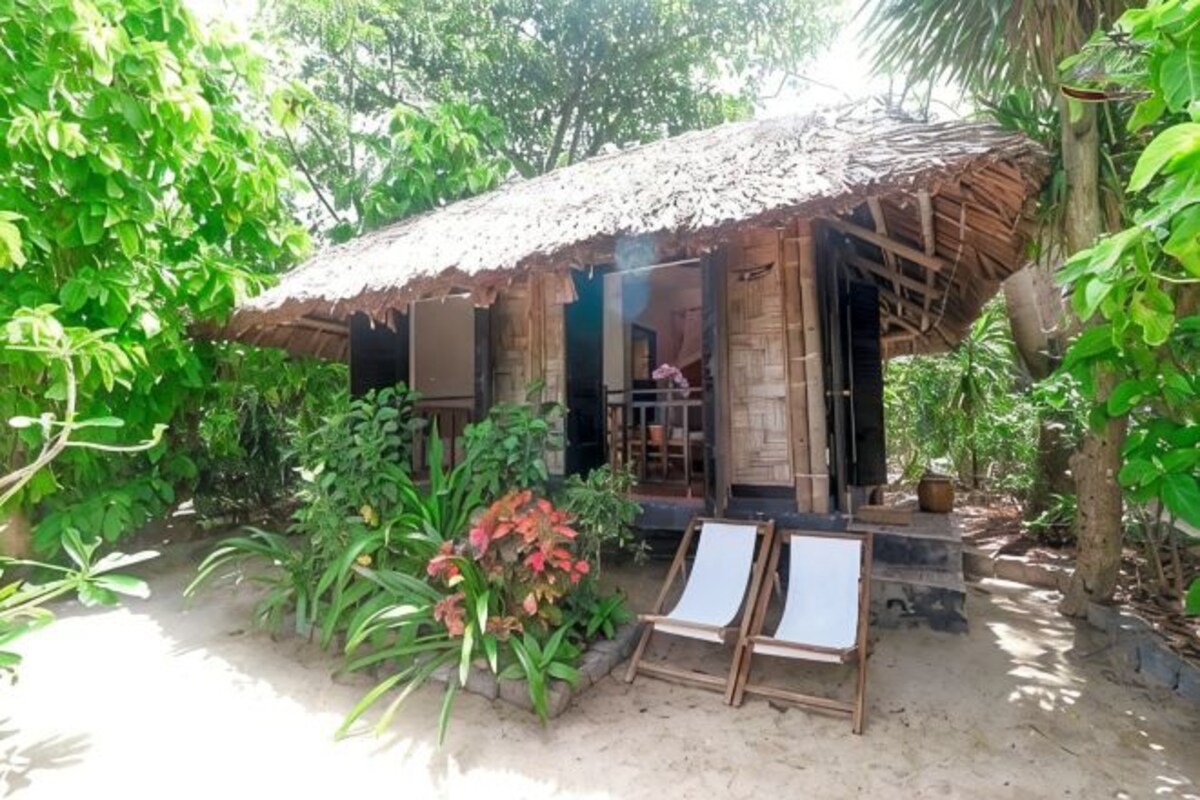 A rustic hut with a thatched roof, surrounded by lush greenery and a small garden area with a wooden bench and a signboard.