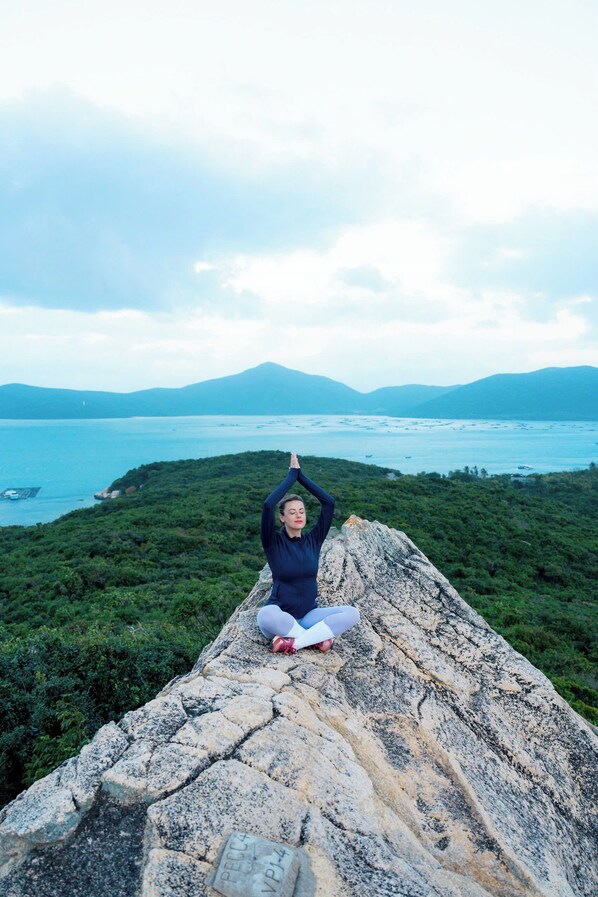 A person meditating on a stone ledge with a view of a coastal landscape and mountains in the distance.