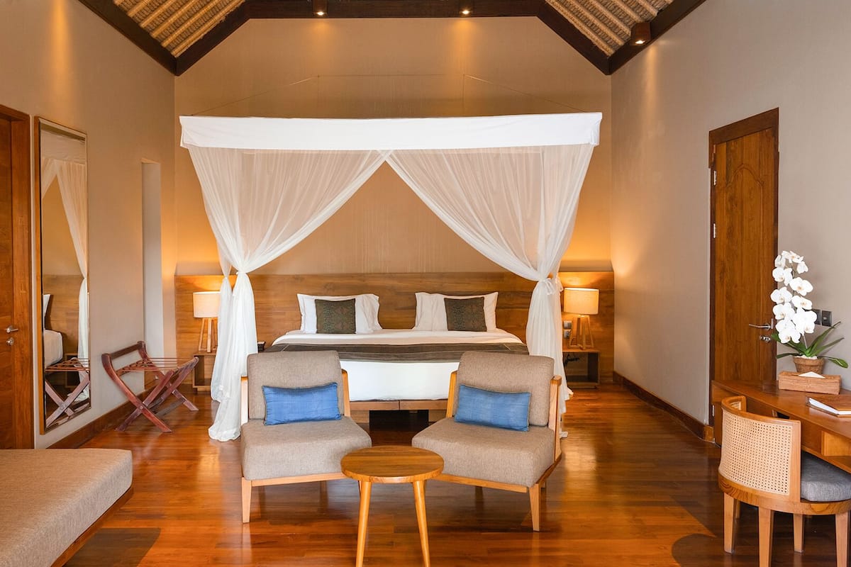 A bedroom with a four-poster canopy bed, a desk with a chair, a small round table, and a mirror.