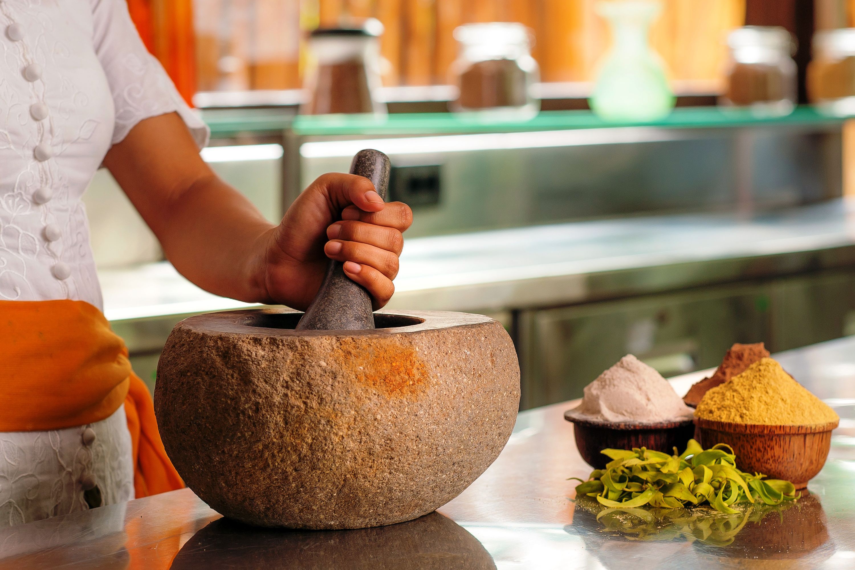A person using a mortar and pestle, with various spices and herbs on the table.