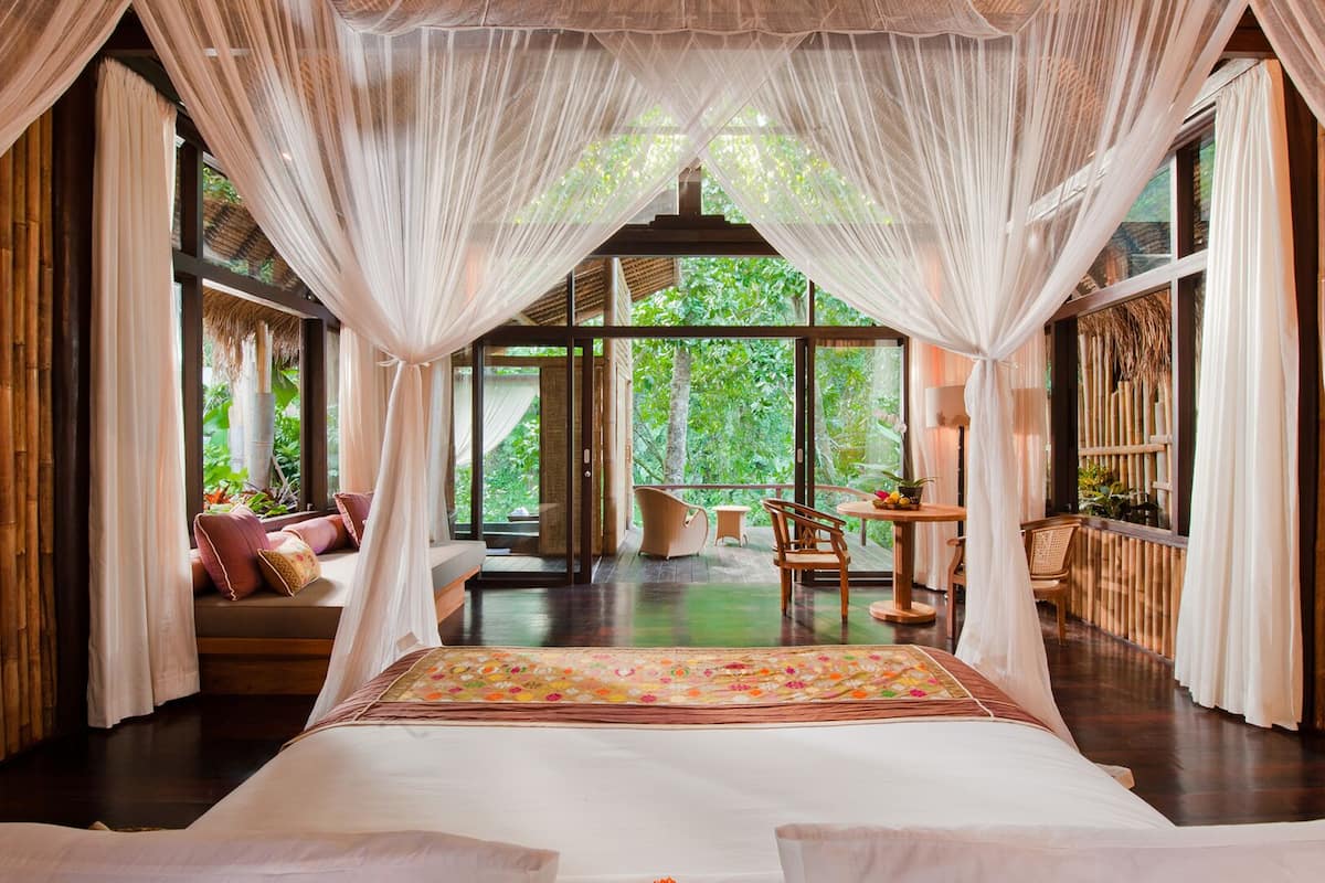 A spacious bedroom with a large bed, a sitting area with a table and chairs, and a view of lush greenery through large windows.