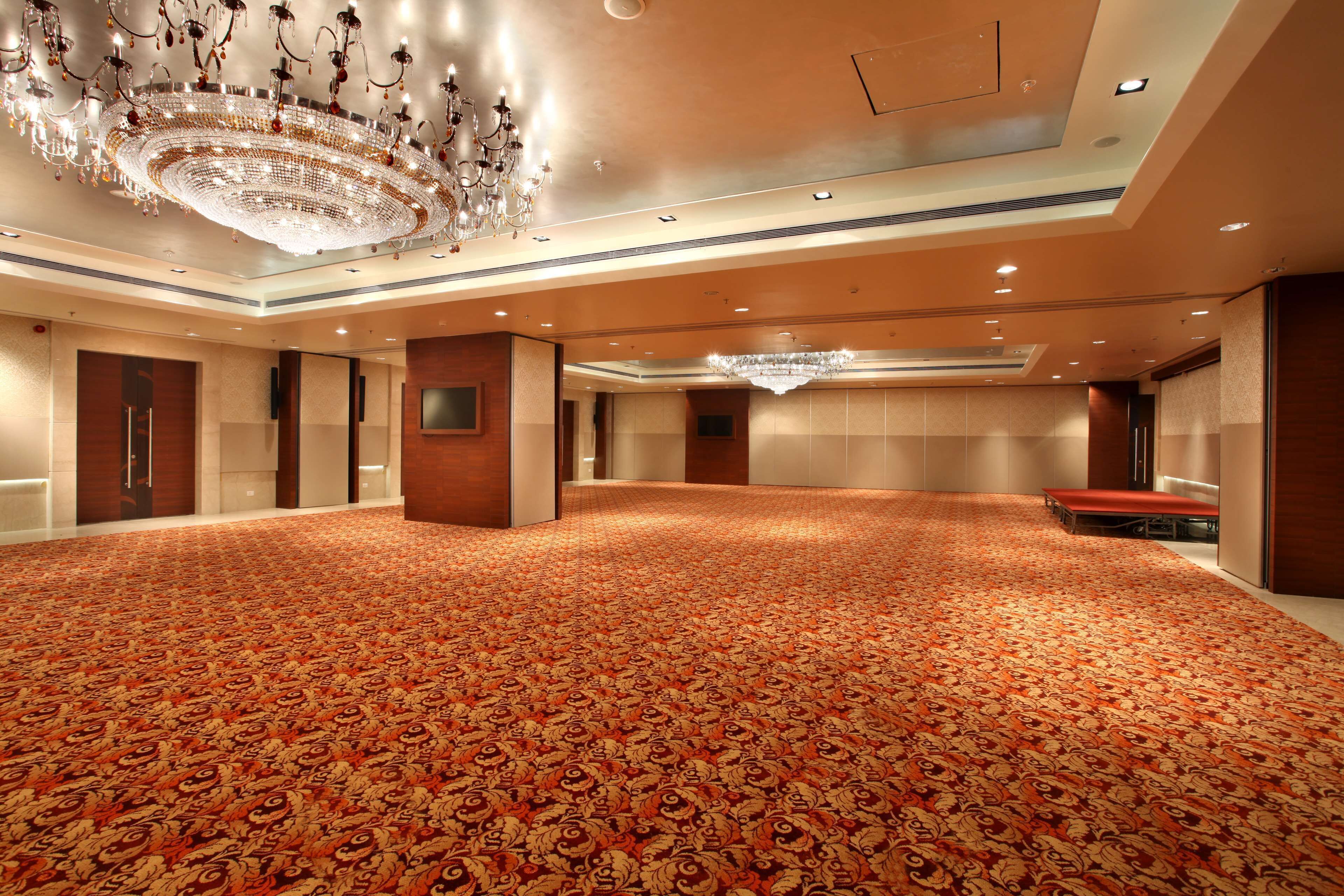 A spacious, carpeted hall with a large chandelier, recessed lighting, and a flat-screen TV mounted on a pillar.