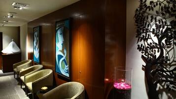 A hotel lobby with a row of armchairs, a decorative wall with a tree design, and a tall pillar with pink lights.