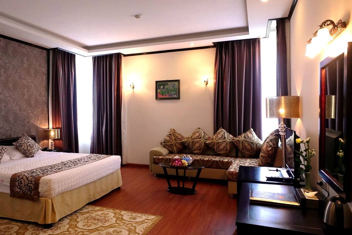 A hotel room with a bed, sofa, coffee table, desk, lamp, and wall art.
