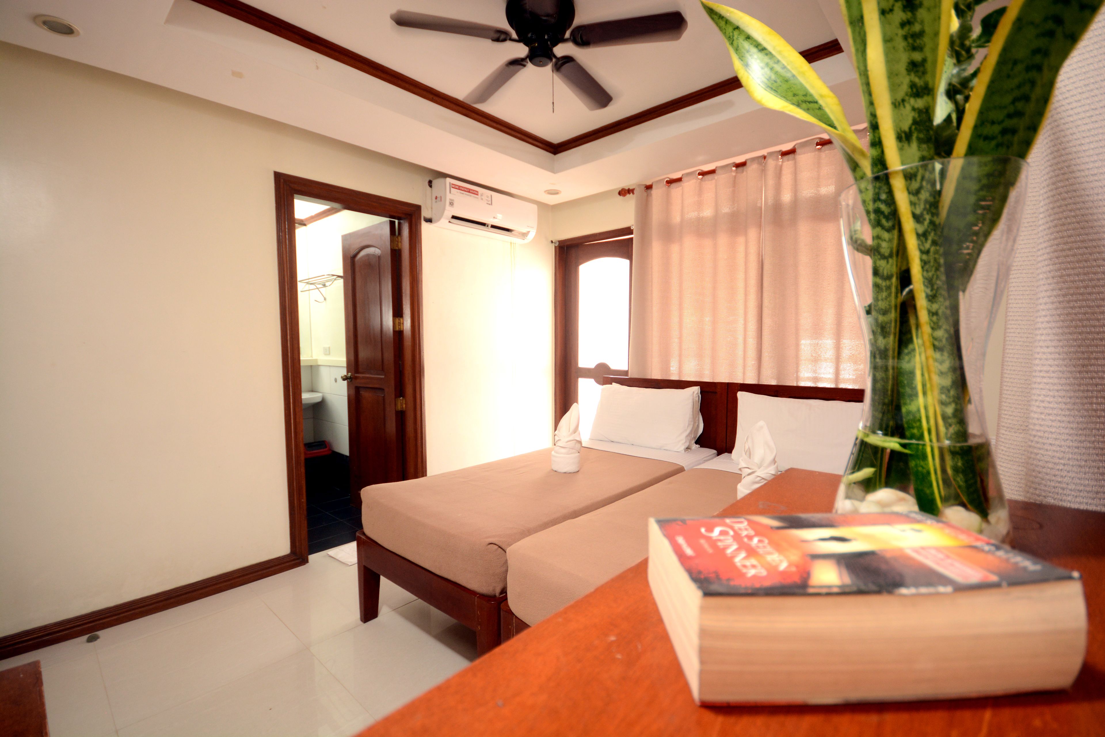 A hotel room with a single bed, a bedside table with a book, a ceiling fan, and a window with curtains.