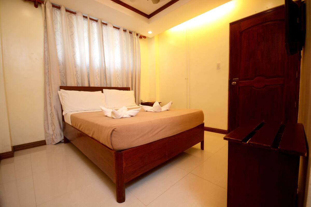 A neatly made bed with white pillows and a brown bedspread in a room with a wooden door and a curtain.