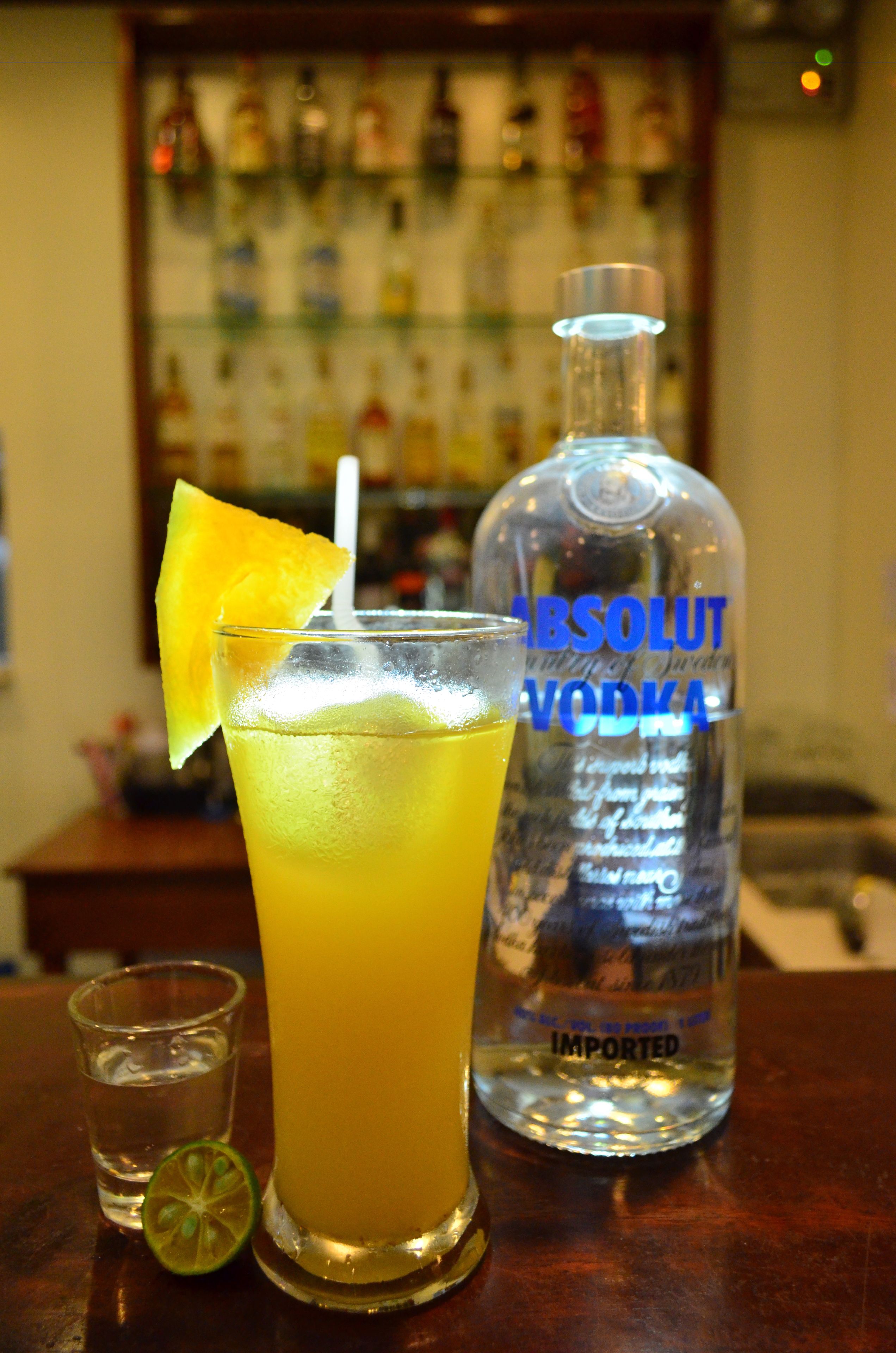 A bar setting with a tall glass of a yellow beverage, a lime wedge, and a bottle of Absolut Vodka.