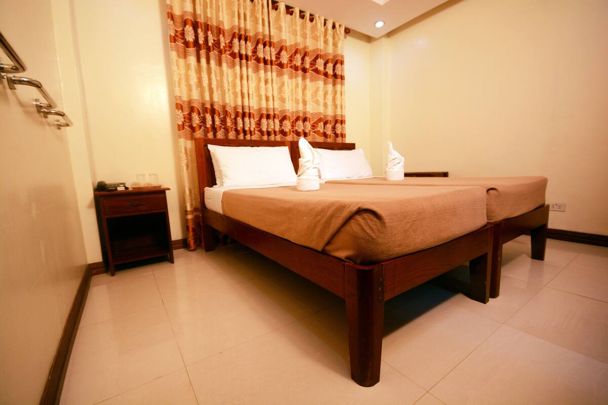 A neatly made bed with a wooden frame and a beige bedspread, a small bedside table with a lamp, and a curtain with a floral pattern.