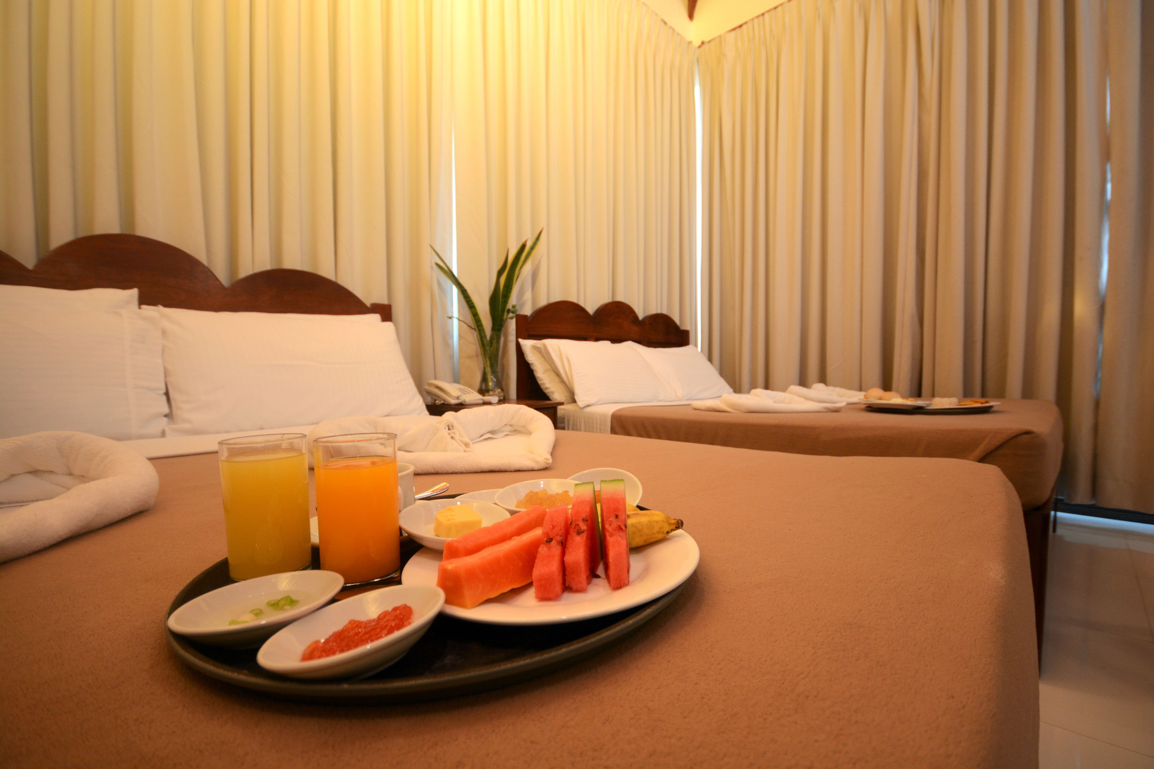 Habitación de hotel con dos camas, una bandeja de desayuno con fruta y jugo, y una planta.
