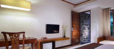 A hotel room with a bed, a desk with a chair, a television, a small refrigerator, and a stone wall.