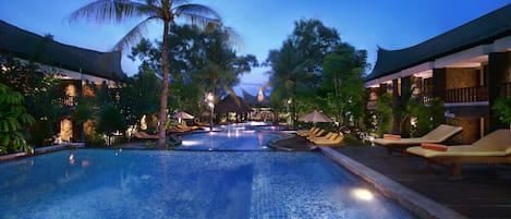A swimming pool with lounge chairs and palm trees, surrounded by buildings with thatched roofs.