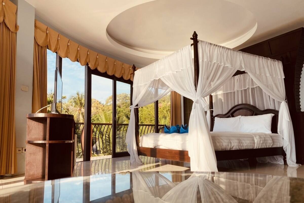 Ein geräumiges Schlafzimmer mit einem Himmelsbett, großen Fenstern mit Vorhängen und Blick auf Palmen.